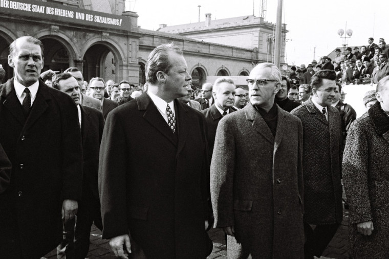 Schwarz-Weiß-Aufnahme von Willy Brandt und Willi Stroph, die von vielen Menschen umringt den Erfurter Hauptbahnhof verlassen. An diesem ist die Schrift zu lesen: „Der Deutsche Staat des Friedensund des Sozialismus!“.