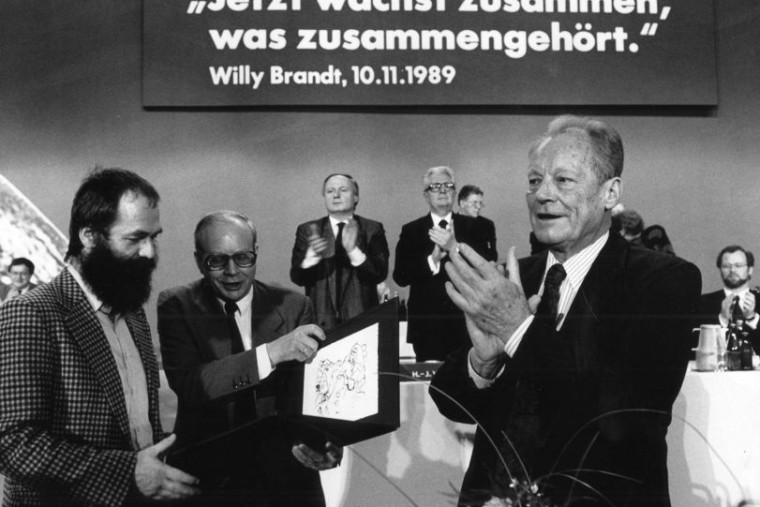 Schwarz-Weiß Aufnahme von Willy Brandt, der von Markus Meckel und Hans Capraro ein Geschenk überreicht bekommt. Im Hintergrund sieht man Oskar Lafontaine und Hans-Jochen Vogel klatschen. Über ihnen der Schriftzug: „Jetzt wächst zusammen, was zusammengehört.“ Willy Brandt. 10.11.1989.