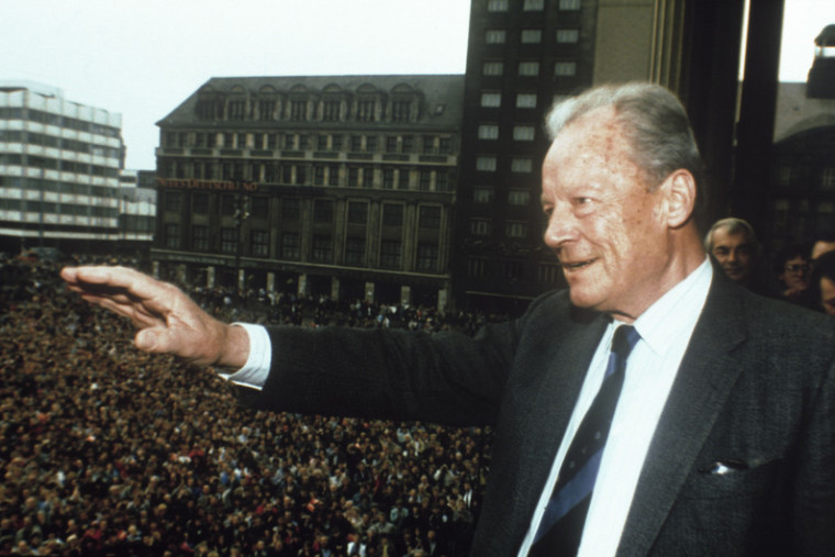Willy Brandt, der auf einem Balkon steht und der Menschenmasse vor Ihm auf dem Platz zuwinkt.
