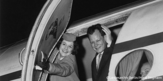 Willy Brandt winkt aus der Tür eines Flugzeuges, während eine Flugbegleiterin diese im Begriff ist zu schließen. Fotografie in Schwarz-Weiß. Öffnet eine Auflistung der Reisen in den Jahren von 1957 bis 1966.