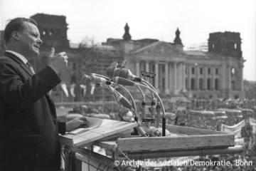 Willy Brandt vor einem Rednerpult bei einer 1. Mai-Kundgebung vor dem Berliner Reichstag. Fotografie in Schwarz-Weiß. Öffnet weitere Informationen zu Brandts Politik in und für Berlin.