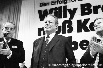 Willy Brandt steht zwischen Herbert Wehner und Helmut Schmidt. Hinter ihm steht halb verdeckt auf der Leinwand: „Den Erfolg fortsetzen. Willy Brandt muß Kanzler bleiben.“ Fotografie in Schwarz-Weiß. Öffnet weitere Informationen zu seiner Zeit als Vorsitzender der SPD.