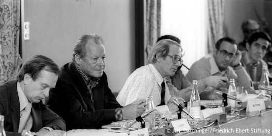 Willy Brandt sitzt neben Fritz Fischer, Göran Ohlin, Jan Pronk und Abdlatif Y. Al-Hamad an einem Tisch bei der Sitzung der Nord-Süd-Kommission. Fotografie in Schwarz-Weiß. Öffnet eine Auflistung der Reisen in den Jahren von 1975 bis 1982.