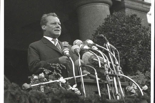 Schwarz-Weiß-Aufnahme Willy Brandts 1961 vor einer Vielzahl von Mikrofonen. Im Hintergrund lässt sich das Rathaus Schöneberg erahnen.