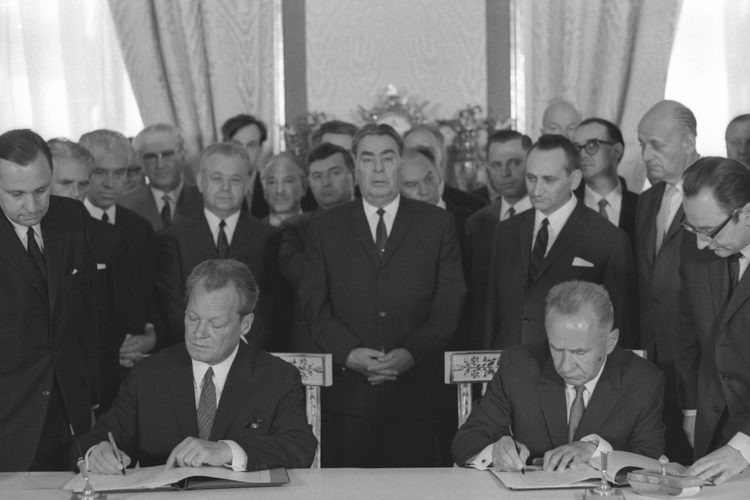 Schwarz-Weiß-Aufnahme Willy Brandts und des sowjetischen Ministerpräsidenten Alexei Kossyhin, die jeweils eine Version des Moskauer Vertrages 1970 unterschreiben. Im Hintergrund sind einige gespannt blickende Politiker zu sehen.