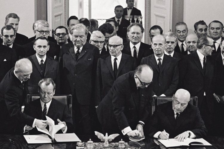 Schwarz-Weiß-Aufnahme Willy Brandts und des polnischen Ministerpräsidenten Józef Cyrankiewicz, die jeweils eine Version des Warschauer Vertrags 1970 unterschreiben. Im Hintergrund sind einige gespannt blickende Politiker zu sehen.