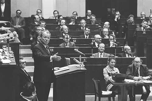 Schwarz-Weiß-Aufnahme Willy Brandts 1973, der seitlich vor einem Rednerpult steht. Im Hintergrund sind Abgeordnete des Europäischen Parlamentes zu sehen, die an ihren Plätzen sitzen.