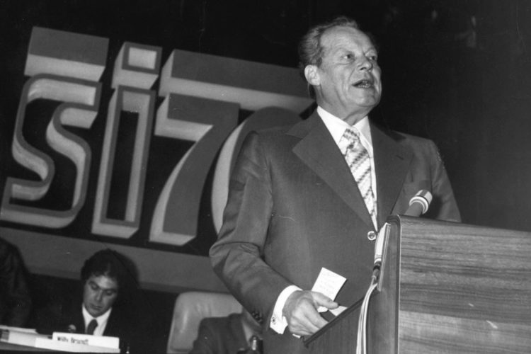 Schwarz-Weiß-Aufnahme Willy Brandts vor einem Rednerpult 1976. Im Hintergrund ist das Logo SI76 zu sehen.