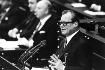 Schwarz-Weiß-Aufnahme von Willy Brandt aus dem Jahre 1969 vor einem Rednerpult im Bonner Bundestag. Links neben ihm ist auf der Regierungsbank Walter Scheel sowie eine Reihe weiterer Minister zu sehen.