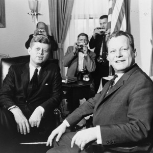 Schwarz-Weiß-Aufnahme von Willy Brandt und John F. Kennedy. Hinter den beiden sind 3 Fotografen zu sehen, die den Finger bereits am Auslöser haben.